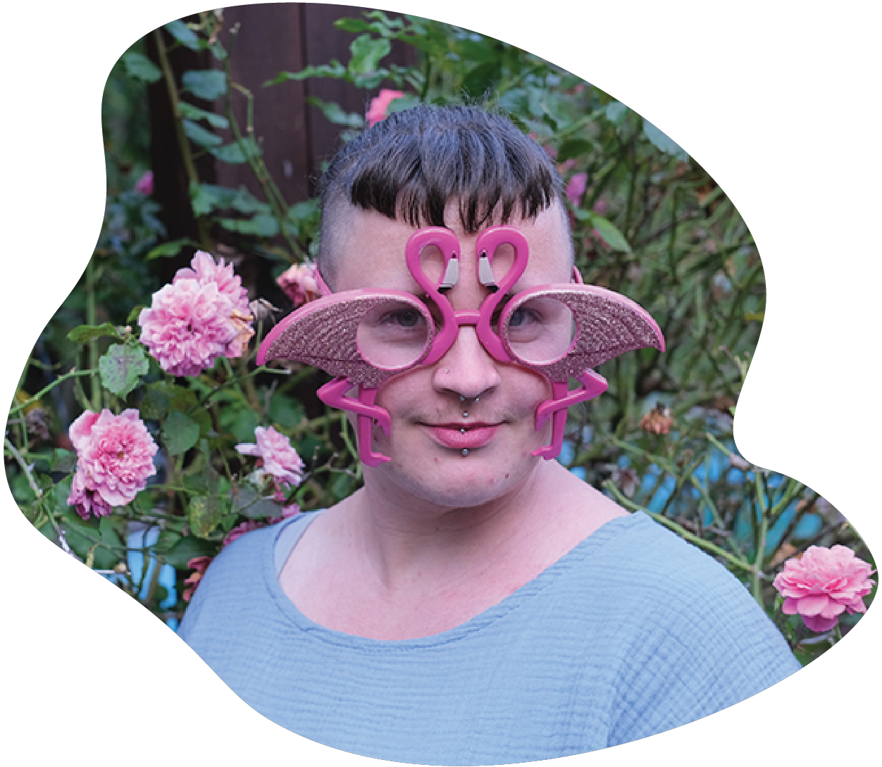 Tristan Marie steht vor einem Rosenbusch, schaut direkt in die Kamera und lächelt leicht. Nin trägt eine pinke, glitzernde Brille in der Form von 2 Flamingos und ein hellblaues Shirt. Tristan Marie ist weiß. Nin hat helle haut und dunkle Haare mit einem kurzen Pony und ausrasierten Seiten.